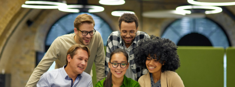 Five coworkers smiling together in an office, looking at a shared screen.
