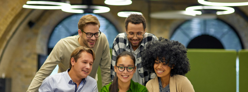 Five coworkers smiling together in an office, looking at a shared screen.